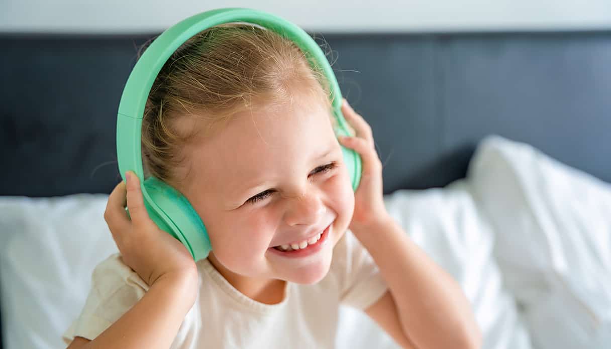 Meisje zit blij op bed, ze luistert naar muziek op haar kindvriendelijke koptelefoon.
