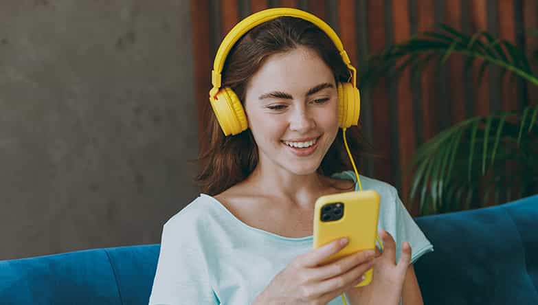 Vrouw zit op de bank te scrollen op haar telefoon en ondertussen maakt ze gebruik van haar bedrade koptelefoon.