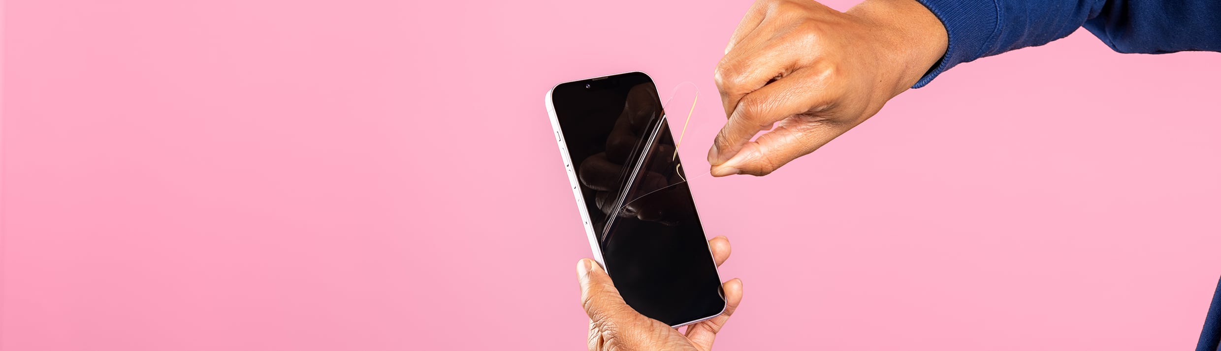 Twee handen die een folie screenprotector verwijderen van een iPhone, de achtergrond is roze.