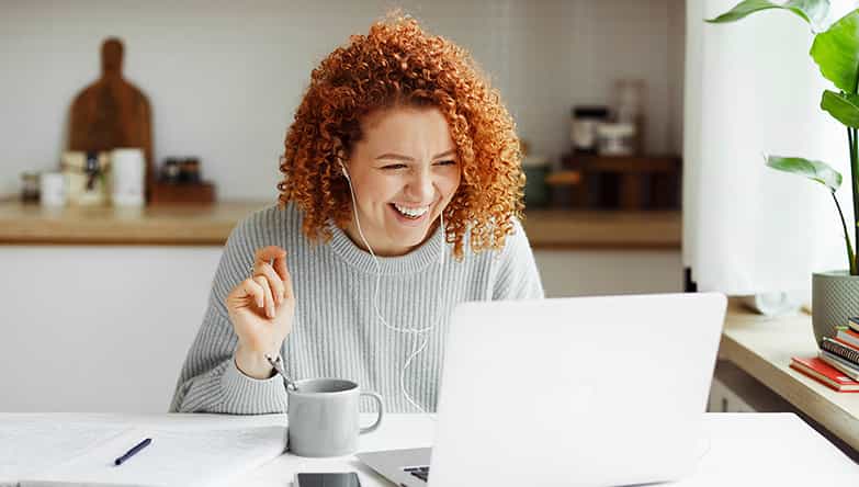Vrouw zit achter haar laptop te lachen naar het scherm erwijl ze oordopjes draagt met draad.