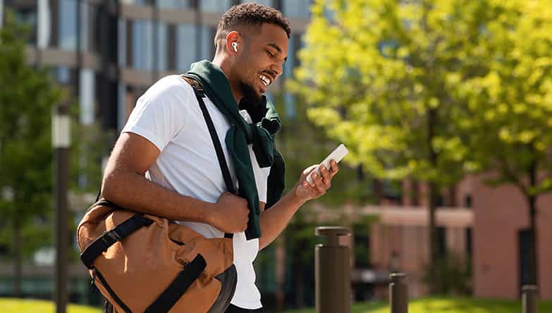Man kijkt lachend naar zijn telefoon, heeft een sporttas om zijn schouder heen en draagt draadloze AirPods in zijn oren.