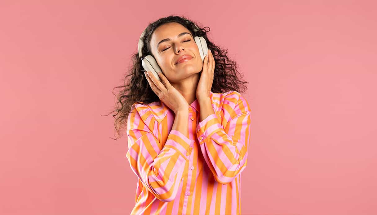 Een vrouw op een roze achtergrond geniet met gesloten ogen van muziek via haar noise-cancelling koptelefoon.