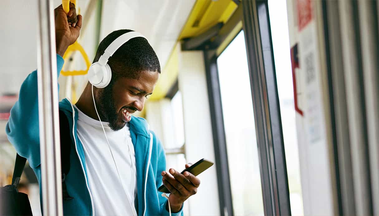 Man staat in de bus lachend naar zijn telefoon te kijken, hij heeft een koptelefoon op met draad.