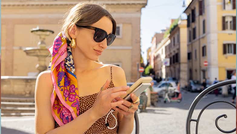Vrouw met zonnebril op zit op een terras gebruik te maken van haar telefoon en powerbank.