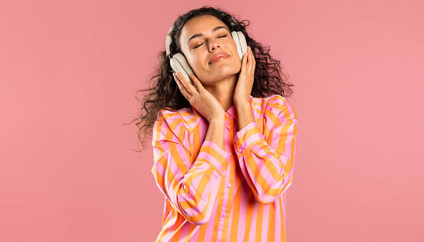 Een vrouw op een roze achtergrond geniet met gesloten ogen van muziek via haar noise-cancelling koptelefoon.