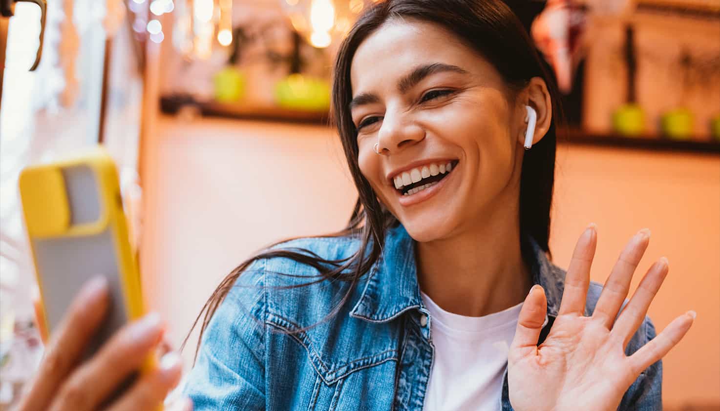 Vrouw zwaait lachend naar haar telefoon, ze is aan het videobellen en heeft haar draadloze oordopjes in.