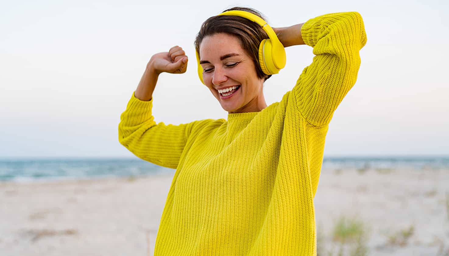 Op het strand staat een lachende vrouw met een felgele koptelefoon met noise cancelling op haar hoofd.
