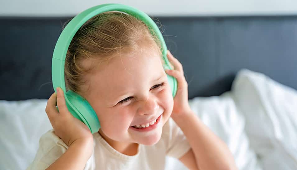 Meisje zit blij op bed, ze luistert naar muziek op haar kindvriendelijke koptelefoon.
