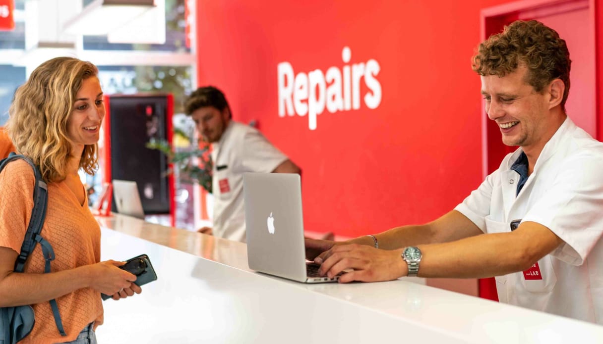 Een vrouw met een laptop aan een witte balie voor laptopreparatie. Aan de andere kant is een reparateur bezig haar laptop werkend te krijgen.