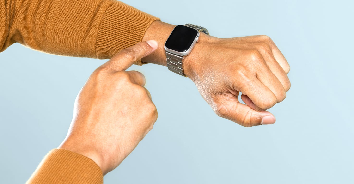 Je ziet een arm met een smartwatch om de pols met een lichtblauwe achtergrond. De trui is geel en de smartwatch wordt bediend met de andere hand.