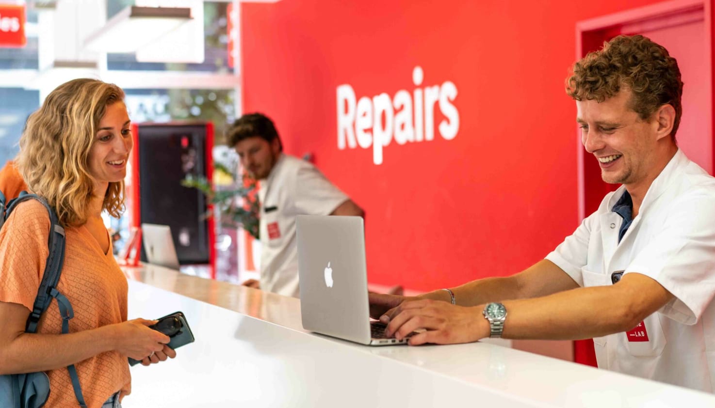 Een vrouw met een laptop aan een witte balie voor laptopreparatie. Aan de andere kant is een reparateur bezig haar laptop werkend te krijgen.